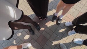 A group of individuals stands in a circle on sunlit paved ground. Only their feet and legs are visible, showcasing a variety of stylish footwear, including black heels, loafers, and white sneakers. The ground consists of gray and brown bricks forming a pattern.